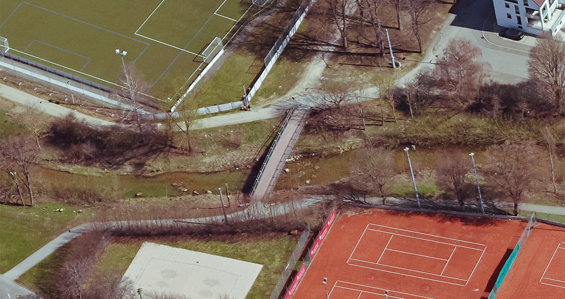 Luftbild von einem Steg über einen Bach, Tennisplätzen und Sportplatz