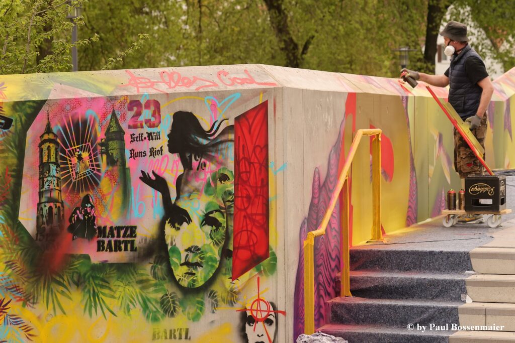 Zu sehen ist ein Mann mit Atemschutzmaske und Spraydose, der an einem farbigen Graffiti an einem Wandabschnitt arbeitet