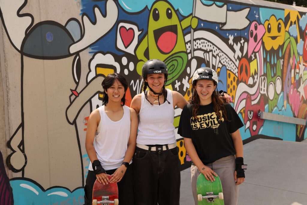 MinhThi, Zoe und Sophia stehen mit ihren Skateboards vor einer bunt besprühten Wand im Aktivpark